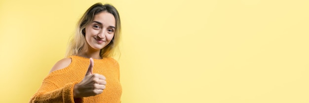 Счастливая молодая кавказская женщина в оранжевом свитере делает большой палец вверх знак и улыбается