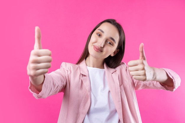 Счастливая молодая кавказская женщина в розовой куртке делает большой палец вверх и улыбается Хорошая работа и уважение