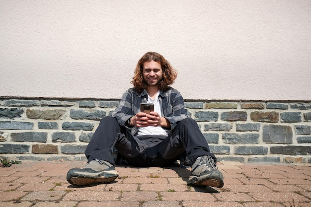 A happy young Caucasian guy sitting in his phone against the wall