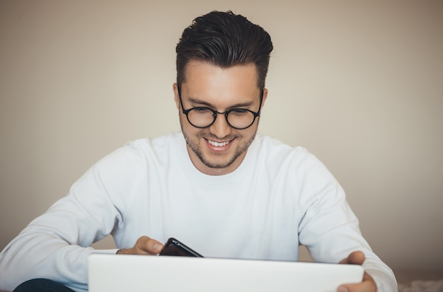眼鏡をかけた幸せな若い白人男性は、ラップトップとモバイルを使用して封鎖中にオンラインで何かを購入しています