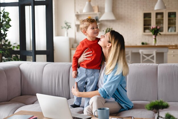 happy young caucasian mother playing and working with her little son kid child at home on freelance remotely single mother on maternity leave