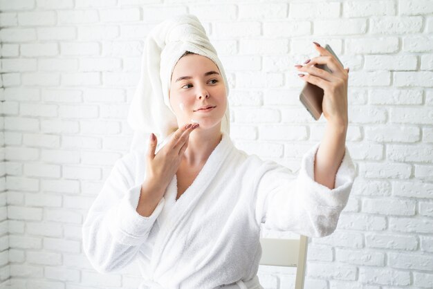 Happy young caucasian woman in white bath towel applying face cream at home doing selfie