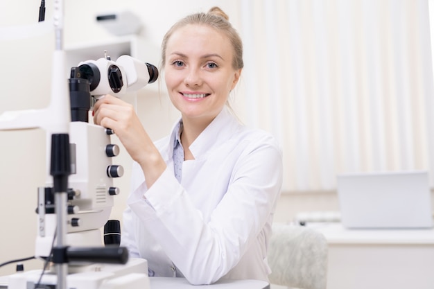 Happy young contemporary ophthalmologist with eye vision check-up equipment looking at you in clinics