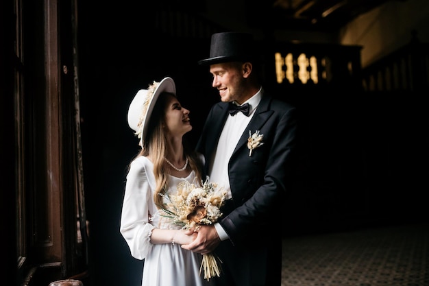 幸せな若いカップルの帽子の花嫁白いウェディングドレスと花の花束を持つ帽子の若い女の子城の花嫁花嫁と新郎