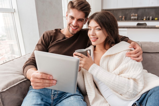 Счастливая молодая пара пьет кофе и вместе дома с помощью планшета на диване