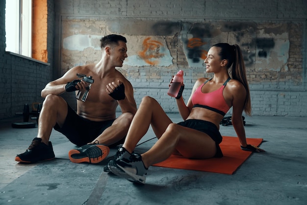 Photo happy young couple drinking water and talking while sitting on exercise mats in gym