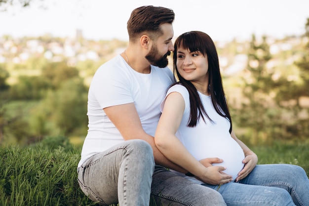 Happy young couple expecting baby, pregnant woman with husband touching belly, sitting on green grass