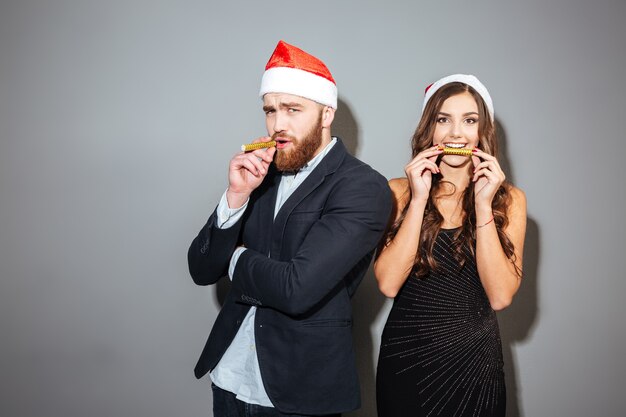 Happy young couple in formal clothes and santa hat celebrating christmas over gray wall