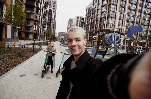 Photo happy young couple has a good time riding e-scooters. fast trip concept. romantic date. young blond man holds camera and takes selfie with his attractive girlfriend.