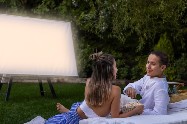 Happy Young Couple Looking To Each Other And Watching A Movie In The Outdoor Summer Cinema With A Bowl Of Popcorn