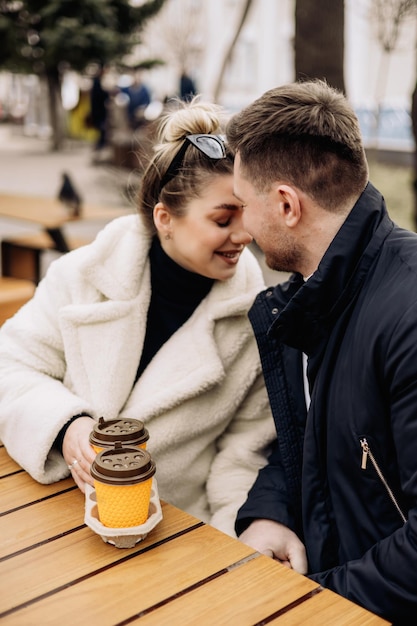 Happy young couple in love in outerwear sitting at a table outside and drinking coffee Relaxing in the open air