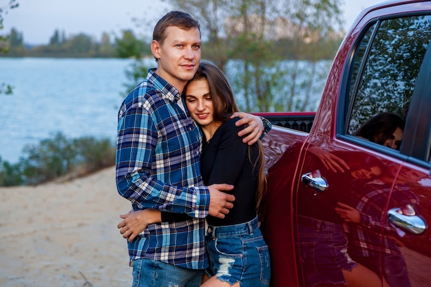 Happy young couple in love smiling and hugging next to red car on the beach near the lake. Young couple hugs near the car.