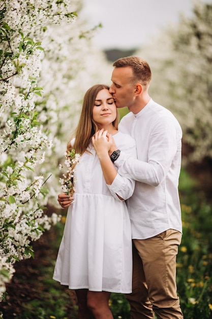 A happy young couple in love stands in a garden of blooming apple trees A man in a white shirt and a girl in a white light dress are walking in a flowering park