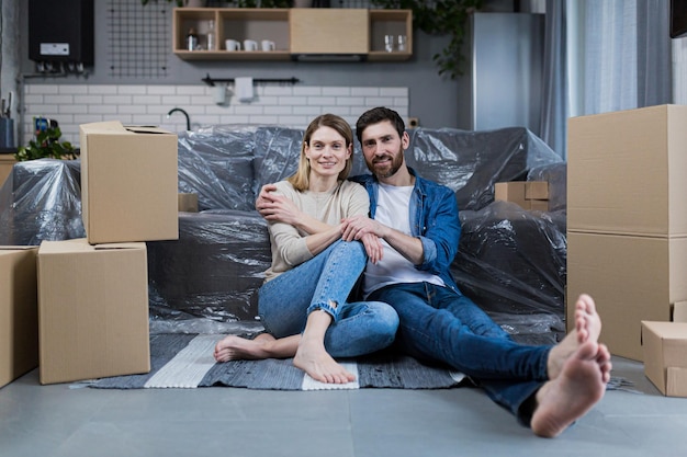 Happy young couple man and woman at home sitting on sofa in new rented apartment near cardboard boxes