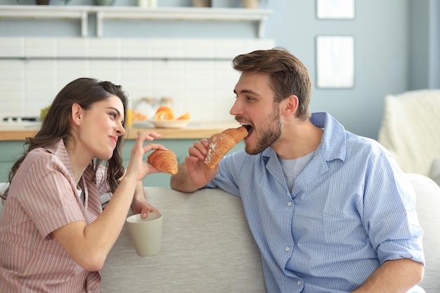 Happy young couple in pajamas in kitchen having breakfast, feeding each other a croissant.