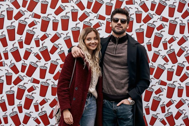 Happy young couple posing at coffe cups background. Young couple in eco fur coats at the street brand wall background. Brand wall designed with coffee cups. Cold season. Young man in sunglasses huggs