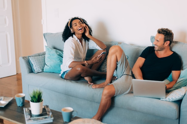 Happy young couple relaxed at home in the couch on the mobile phone listening to music and on the computer