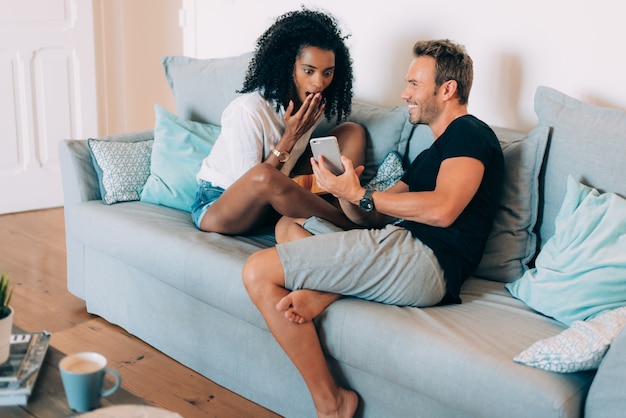 Счастливая молодая пара отдыхает дома на диване, читает книгу и смотрит на мобильный телефон