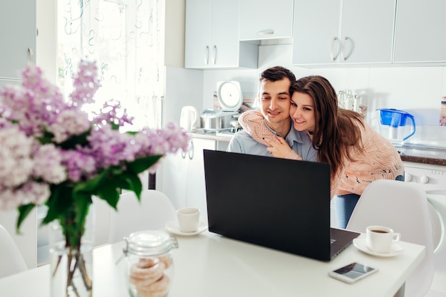 Happy young couple self isolated at home using laptop drinking coffee