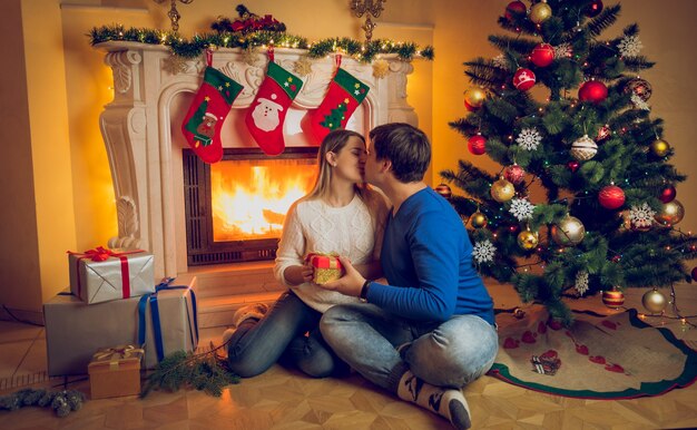 暖炉のそばに座って、クリスマスイブにキスする幸せな若いカップル