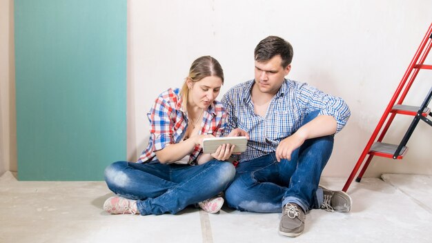 Happy young couple sitting on floor and choosing furniture for their new house under renovation.