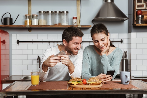 Happy young couple sitting at the kitchen during breakfast at home, using mobile phone