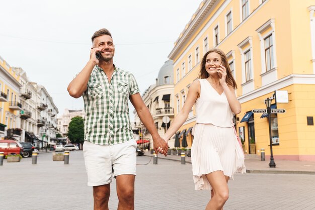 happy young couple in summer clothes holding hands together and talking on smartphones while walking through city street