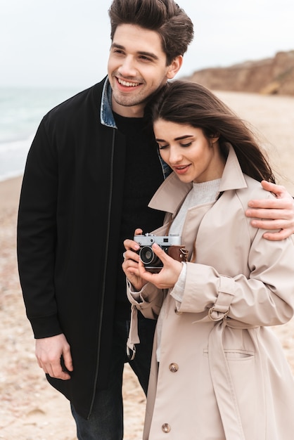 Happy young couple wearing autumn coats spending time together at the seaside, taking pictures with photo camera