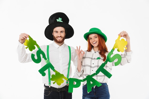 Happy young couple wearing costumes, celebrating StPatrick 's Day isolated over white wall, having fun together, holding decoration