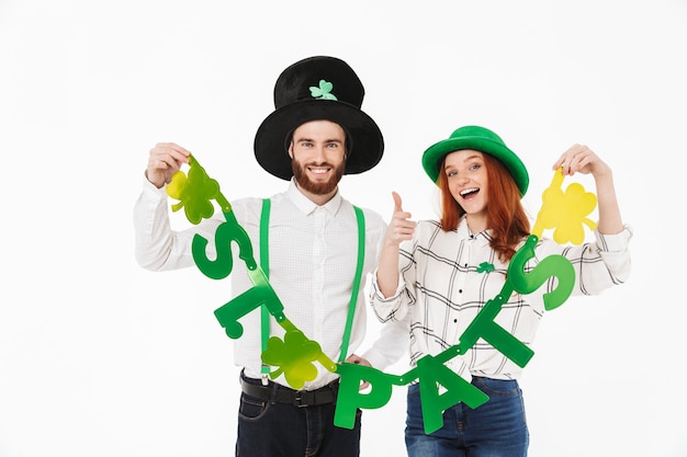 Giovani coppie felici che indossano costumi, celebrando il giorno di stpatrick isolato sopra il muro bianco, divertendosi insieme, tenendo la decorazione