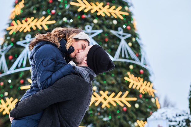 Счастливая молодая пара в теплой одежде стоит возле городской рождественской елки, наслаждаясь проведением времени вместе