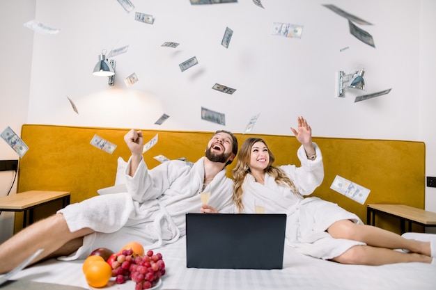 Happy young couple in white bathrobes throwing currency notes in air, while lying on the bed in modern bedroom or hotel.