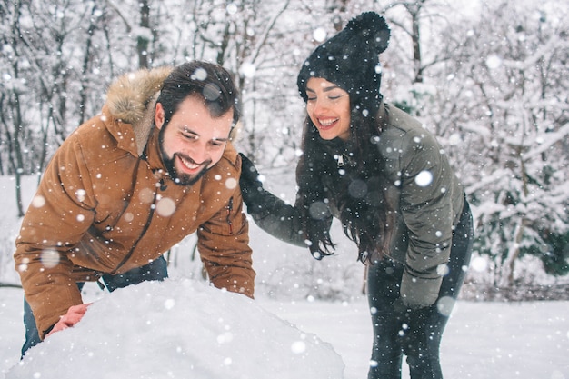 Счастливая молодая пара в зимний период. Семья на открытом воздухе. мужчина и женщина смотрит вверх и смеется. Любовь, веселье, сезон и люди - гуляют в зимнем парке. Создание снеговика.