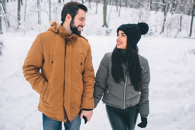 冬の幸せな若いカップル。家族のアウトドア。男と女が上向きに見て笑っています。愛、楽しさ、季節、そして人々-冬の公園を歩いています。お互いの手を立てて