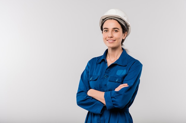 Happy young cross-armed female engineer in blue workwear and hardhat standing in isolation