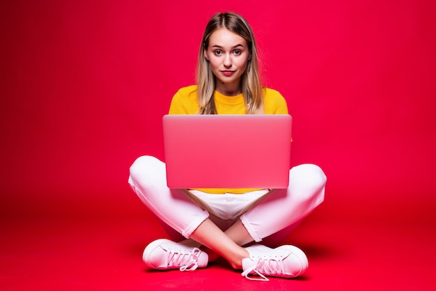 Happy young curly beautiful woman sitting on the floor with crossed legs and using laptop on red wall.