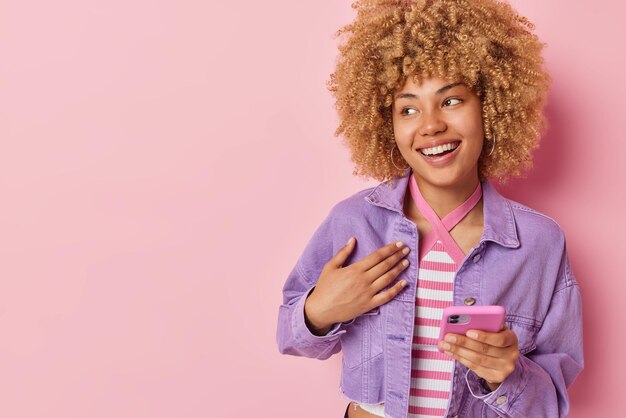 Happy young curly woman uses mobile phone reads good news makes\
shopping via online store app dressed in purple jacket looks away\
smiles positively isolated over pink background blank space