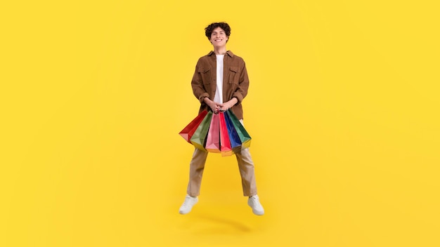 Happy young customer guy holding shopping bags on yellow background