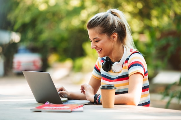 Happy young cute girl student sitting in park using laptop computer.