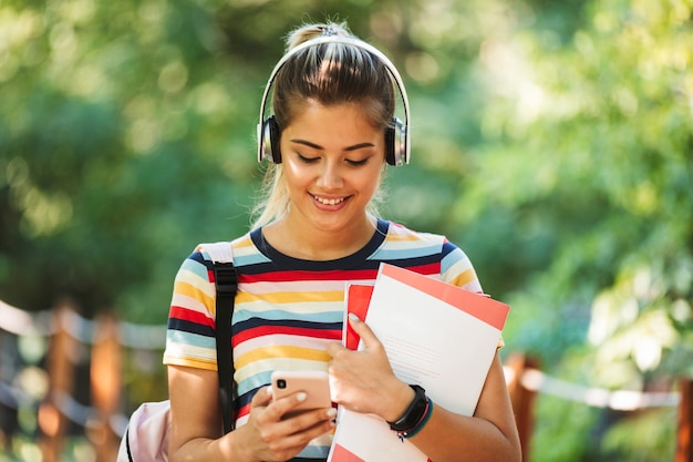 Happy young cute girl student walking in park with backpack using mobile phone listening music.