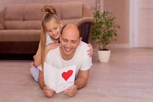 Счастливый молодой папа держит открытку от маленьких веселых дочерей в день отцов во время домашнего праздника
