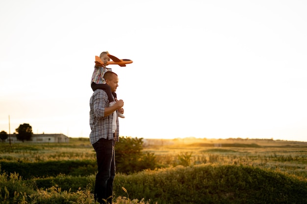 Happy young dad with little daughter on his shoulders in a field at sunset in summer A small family of a caring father and a foster girl on a walk with a toy time togetherplace for text