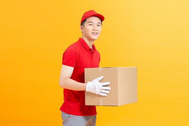 A happy young delivery man in red cap standing with parcel post box isolated over yellow background