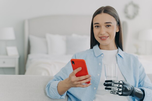 Happy young disabled girl holding smartphone and glass of water using bionic prosthetic arm