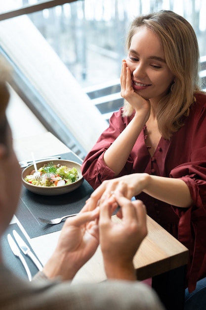 Happy young elegant blond woman looking at her boyfriend putting engagement ring on her finger while both sitting by served table