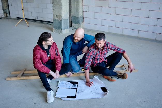 Счастливые молодые инженеры сидят на полу внутри недостроенного здания и обсуждают точки эскиза на чертеже на рабочей встрече