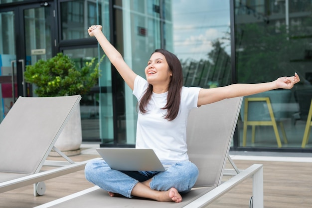 Happy young entrepreneur woman sitting on tanning bed beside pool and using laptop computer