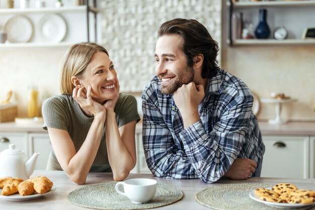 Happy young european female and male with stubble look at each other drink tea in modern kitchen