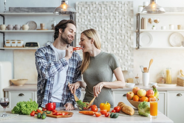 Happy young european husband with stubble feeds woman lady prepares salad for dinner