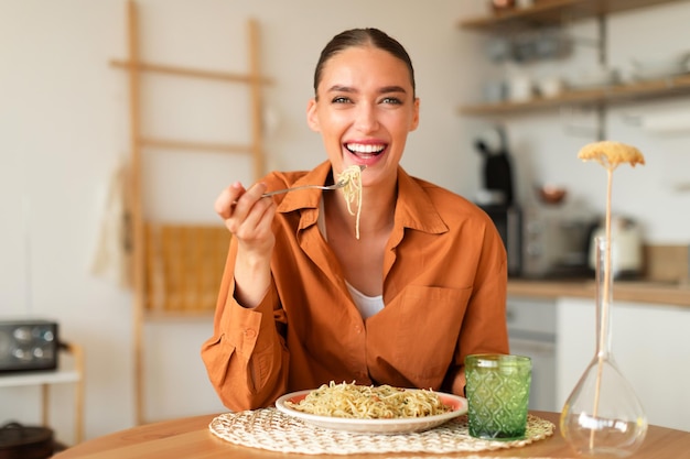 Happy young european lady eating homemade italian pasta enjoying tasty lunch sitting in kitchen